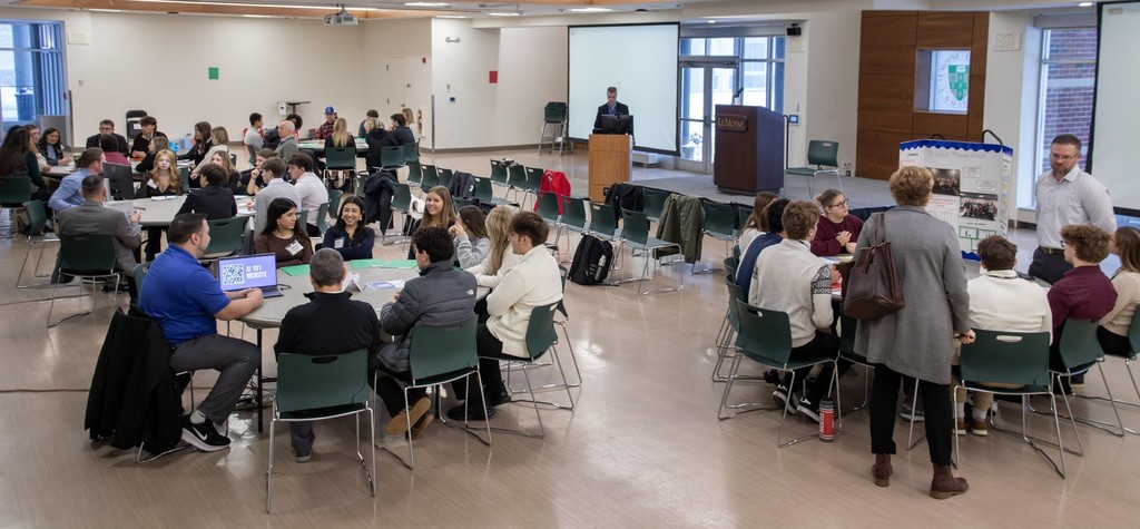A wide view of a large workshop room with multiple tables of high school students participating in small-group discussions. Presenters and facilitators move between tables, and a podium and screen are visible at the front of the room.