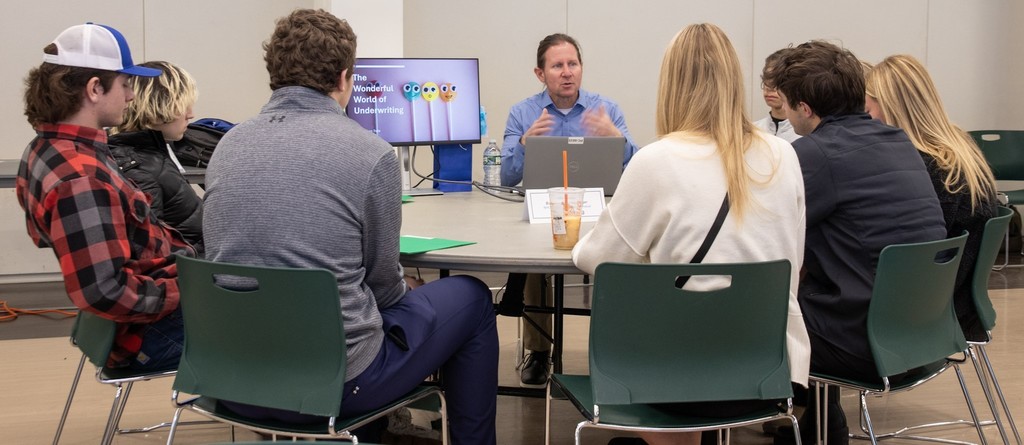A group of high school students sits around a table listening to a presenter during a workshop. The presenter speaks while gesturing, and a screen behind him displays a presentation about underwriting.