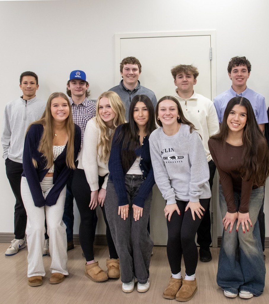 A group of high school students poses together for a photo indoors following a workshop. The students stand in two rows and smile toward the camera.