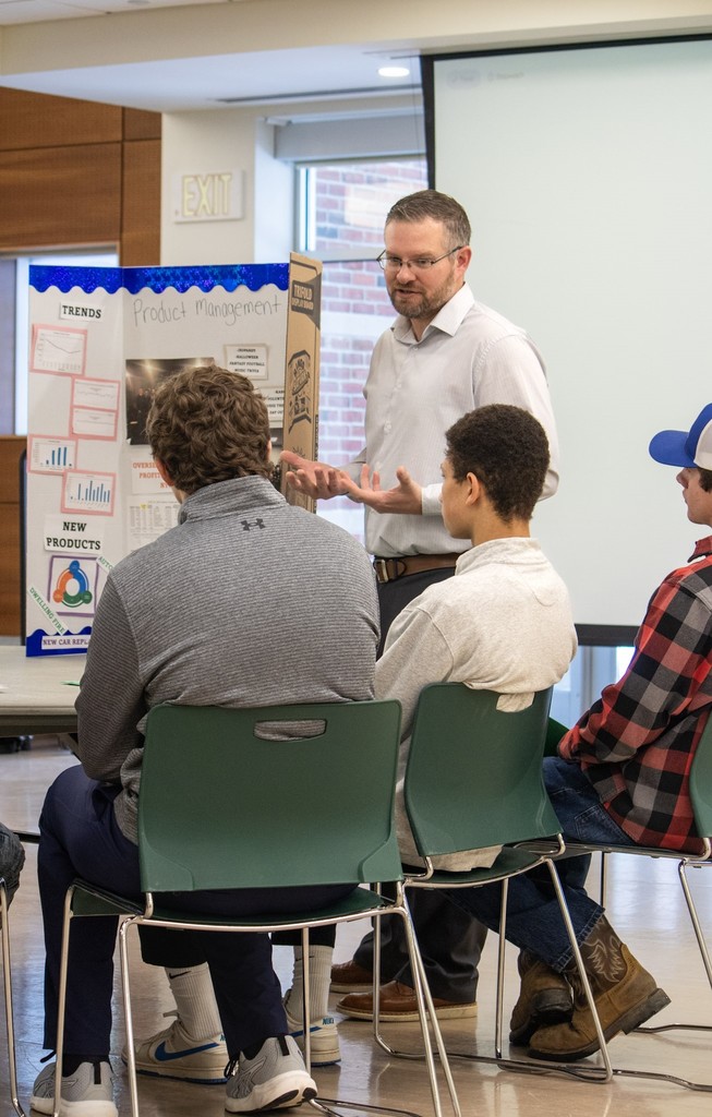 An industry professional stands and speaks to a small group of seated high school students during a workshop. A presentation board about product management is visible nearby as students listen and ask questions.