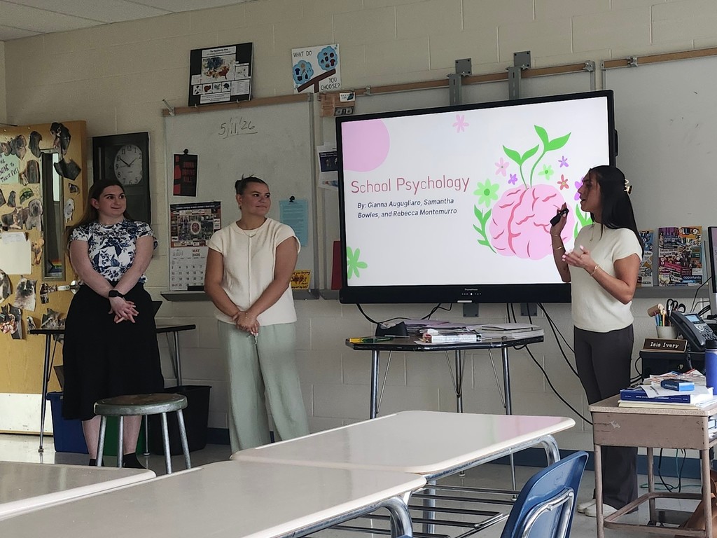 Three SUNY Cortland psychology students stand at the front of a high school classroom presenting about school psychology beside a large interactive display showing their presentation slide.
