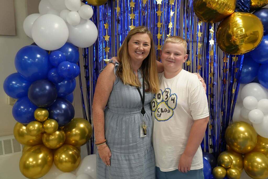 A fourth grade student smiles with his mom in front of a blue, gold, and white balloon display during Lansing CSD's Fourth Grade Moving Up Ceremony.