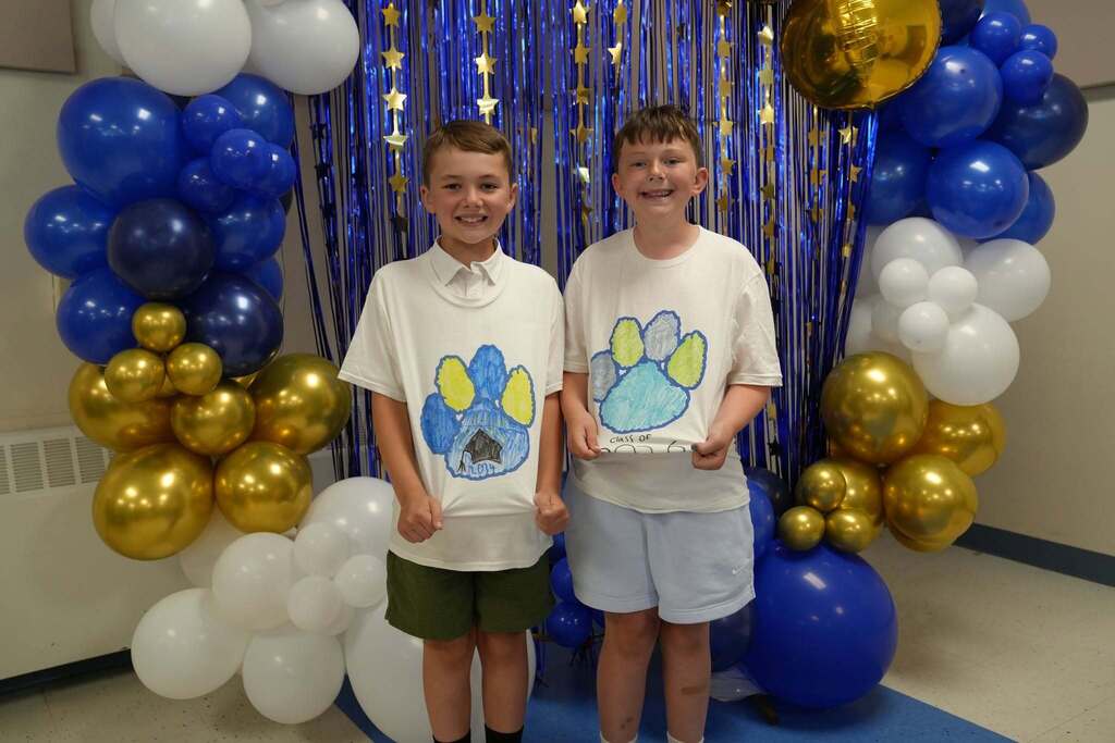 Two fourth grade students wearing custom Class of 2034 T-shirts smile together in front of a blue and gold graduation-themed backdrop.
