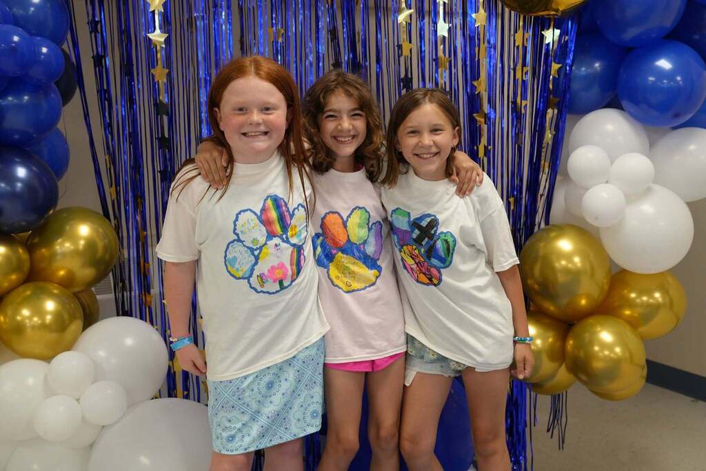 Three fourth grade friends wearing custom Class of 2034 T-shirts smile with their arms around each other in front of a blue and gold celebration backdrop.