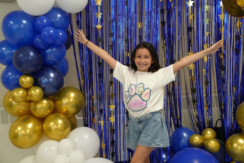 A smiling fourth grade student wearing a custom Class of 2034 T-shirt poses with arms outstretched in front of a blue and gold celebration backdrop.