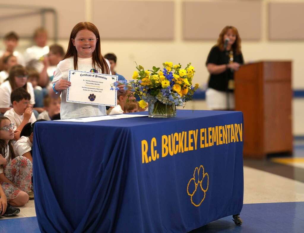 A fourth grade student proudly holds a promotion certificate beside a table decorated with blue and yellow flowers during the Moving Up Ceremony. A staff member speaks at the podium in the background.