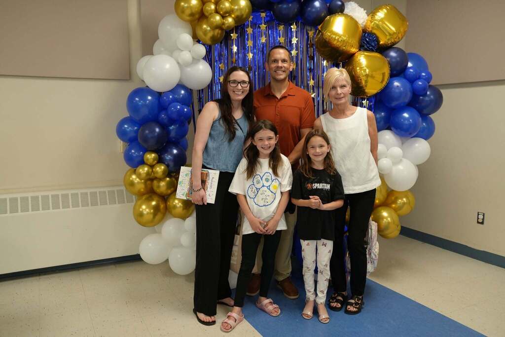 A fourth grade student poses with family members in front of a blue, gold, and white balloon display after the Moving Up Ceremony.