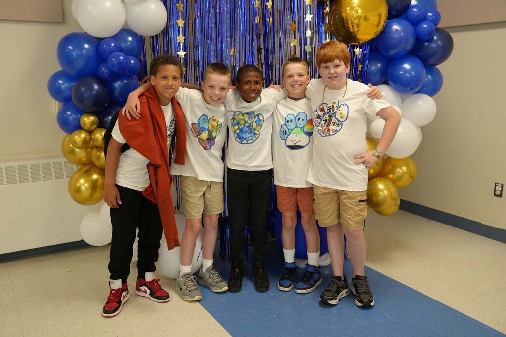 Five fourth grade students wearing custom Class of 2034 T-shirts smile together in front of a blue, gold, and white balloon display.