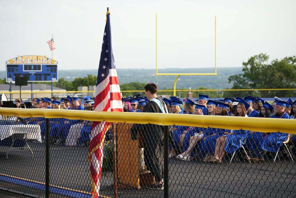 Superintendent Pettograsso addresses the Lansing High School Class of 2026 from a podium during the outdoor commencement ceremony. Graduates in blue caps and gowns are seated behind the speaker, with the American flag and football field visible.