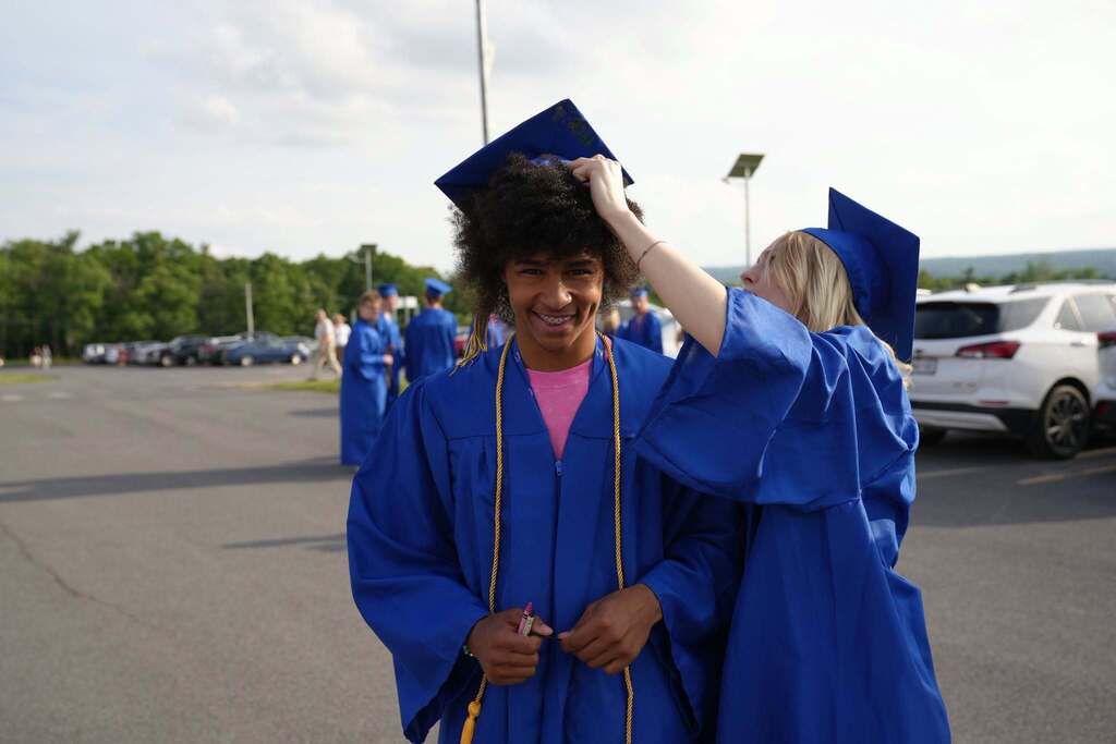 One Lansing High School graduate adjusts another graduate's cap while they smile together before the commencement ceremony in the school parking lot.