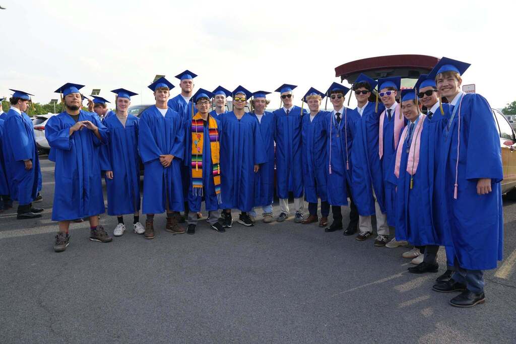 A large group of Lansing High School graduates in blue caps and gowns gather for a class photo before graduation. Several students wear honor cords, stoles, medals, and sunglasses while smiling together.