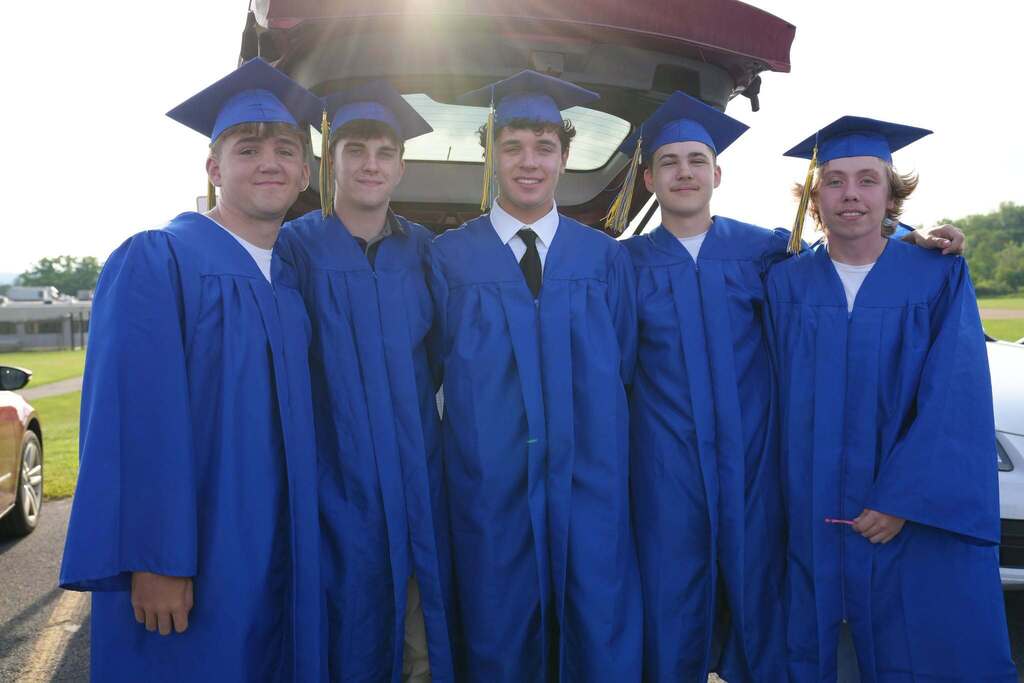 Five Lansing High School graduates in blue caps and gowns pose together in a parking lot before the graduation ceremony, smiling in front of an open vehicle hatch.