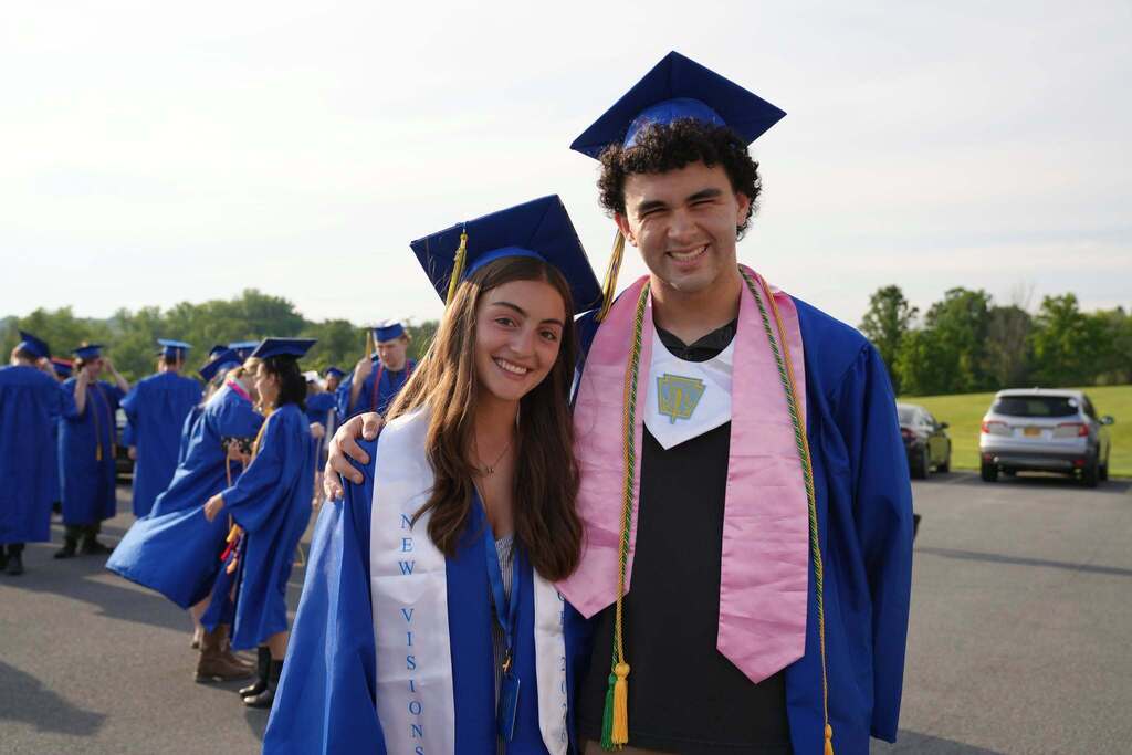 Two Lansing High School graduates smile with their arms around each other before graduation. One wears a New Visions stole, and the other wears a pink honor stole and graduation cords.