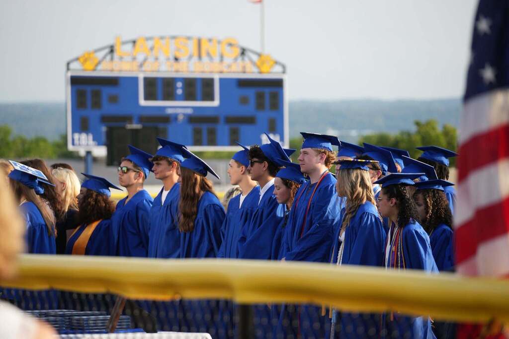 Graduates in blue caps and gowns stand together during the Lansing High School Class of 2026 commencement ceremony, with the Lansing scoreboard visible in the background.
