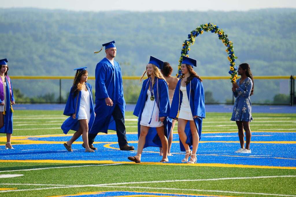 Lansing High School graduates in blue caps and gowns walk across the football field during commencement toward a blue and gold floral arch, with rolling hills in the background.