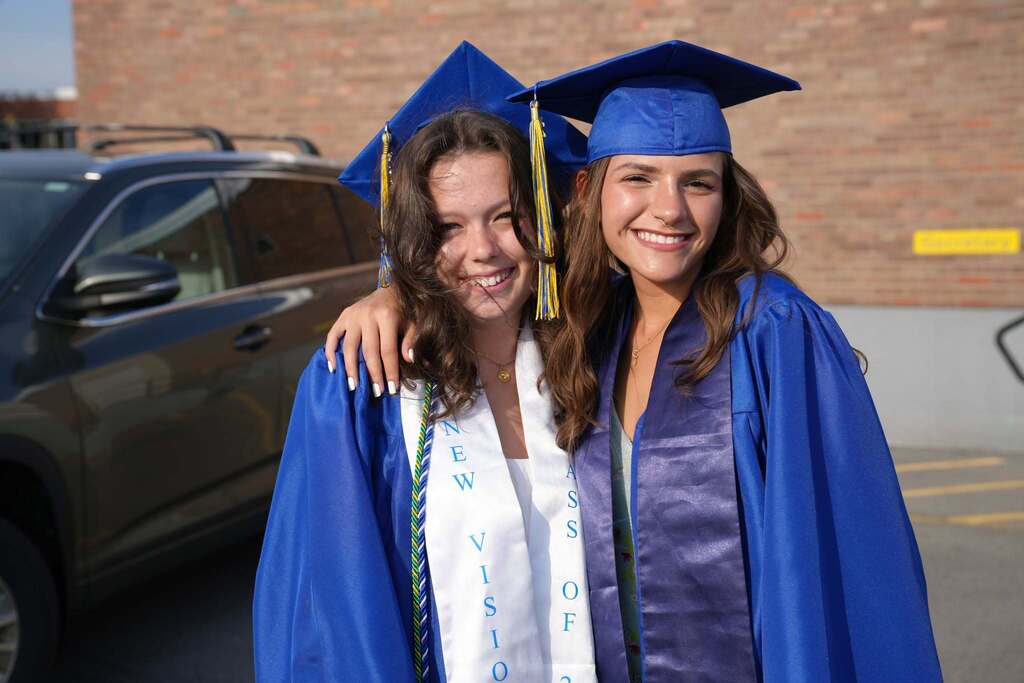 Two Lansing High School graduates in blue caps and gowns smile with their arms around each other before graduation. One wears a New Visions stole, and the other wears an honor stole.