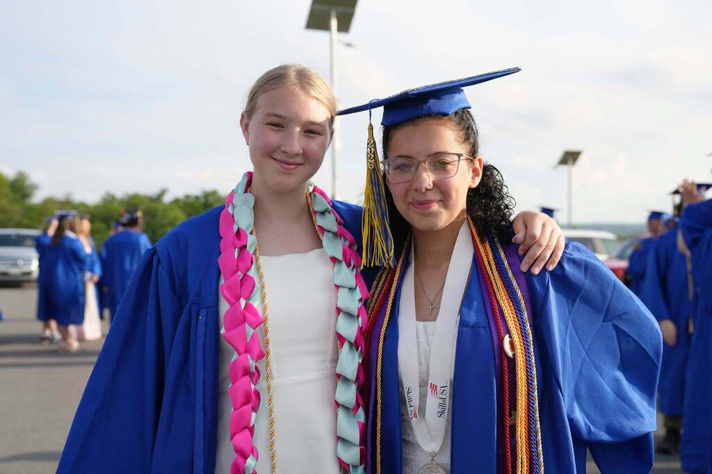 Two Lansing High School graduates in blue caps and gowns smile with their arms around each other before graduation. One wears colorful paper leis, and the other wears honor cords, a medal, and a graduation stole.