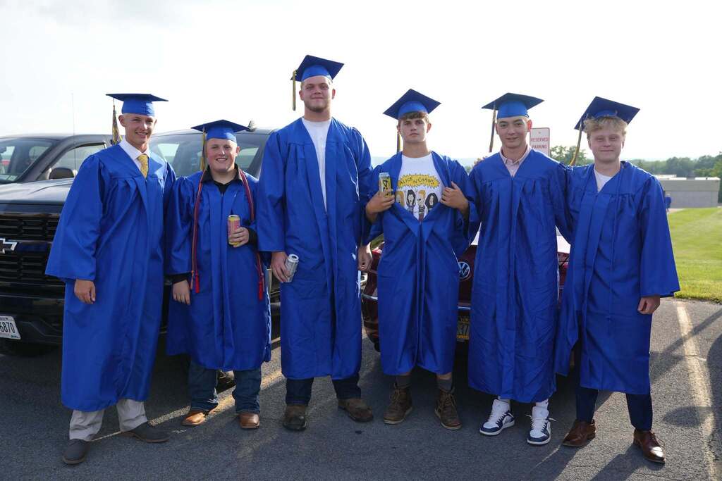 Six Lansing High School graduates in blue caps and gowns pose together before the graduation ceremony, smiling in a parking lot. One graduate playfully opens their gown to reveal a graphic T-shirt.