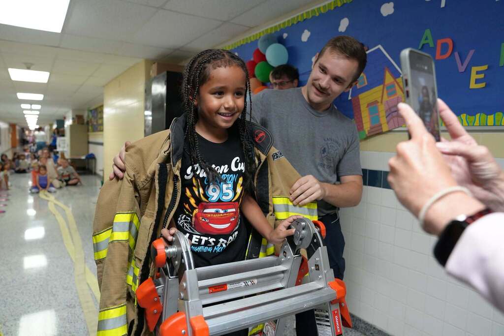 A young student smiling while wearing a heavy firefighter jacket and holding a small ladder with help from a firefighter.