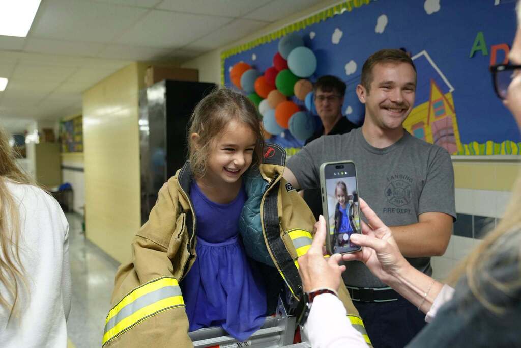 A young student smiling while wearing a heavy firefighter jacket and holding a small ladder with help from a firefighter.