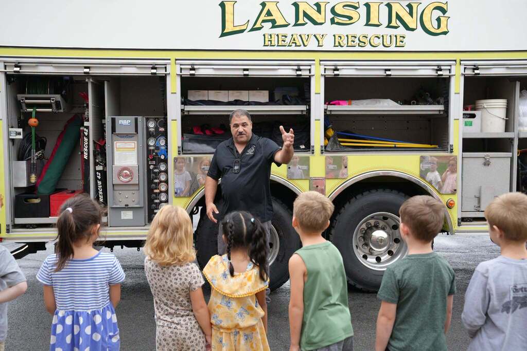 Kindergarten students gather outside listening attentively to a firefighter giving a demonstration in front of a yellow Lansing Heavy Rescue truck