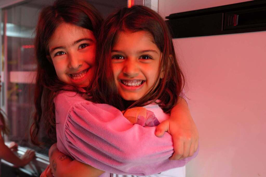Two young girls smiling and hugging inside a fire truck illuminated with red emergency lights.