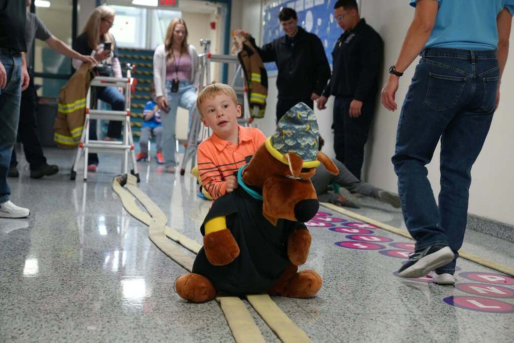 A young boy smiling as carries a plush dog toy to safety and scoots along a flat unrolled fire hose laid out in the hallway.