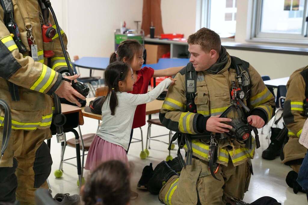 firefighter kneeling in full gear in a classroom while young students reach out to gently touch his turnout jacket.