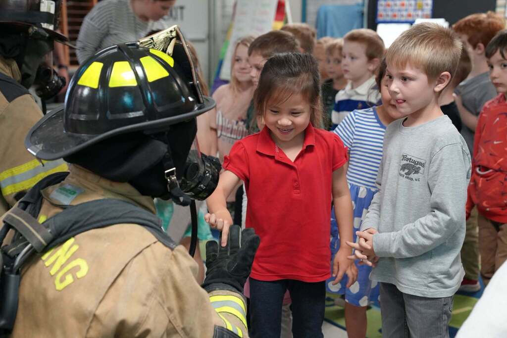Kindergarteners standing in line to high-five and touch the gloved hand of a firefighter wearing full protective gear and a helmet.