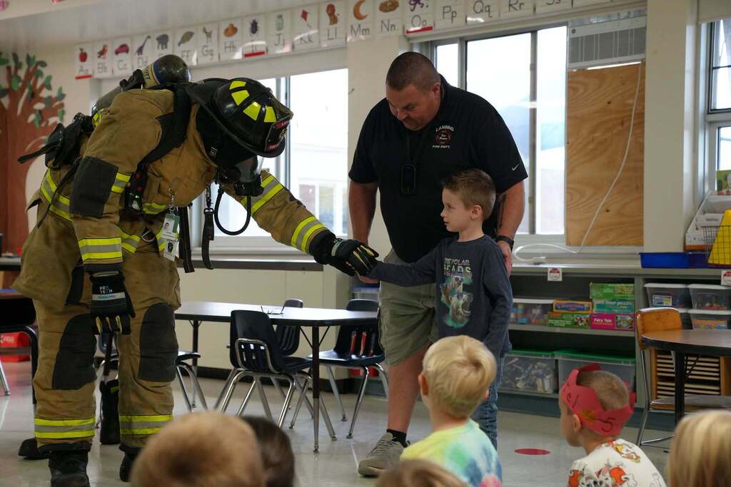 A firefighter in full turnout gear and helmet bending down to hold hands with a young student in front of the class.