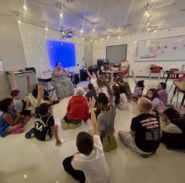 Students sitting on the floor in their classroom raising their hand for the teacher