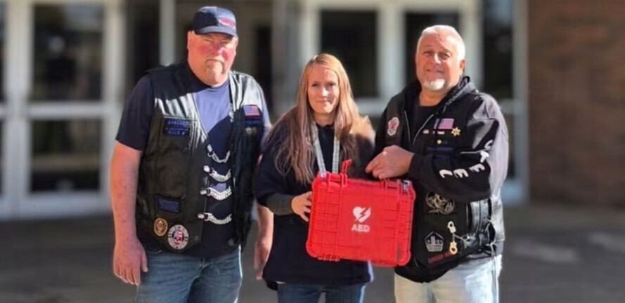 Two gentlemen with the Steel Patriots motorcycle club and LHS athletic secretary pose with an AED