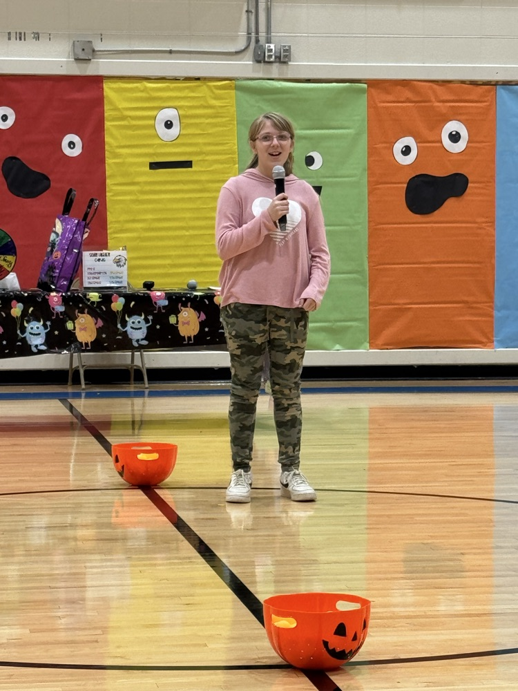 A blonde girl in a pink hoodie sings into the microphone in an elementary school gym.