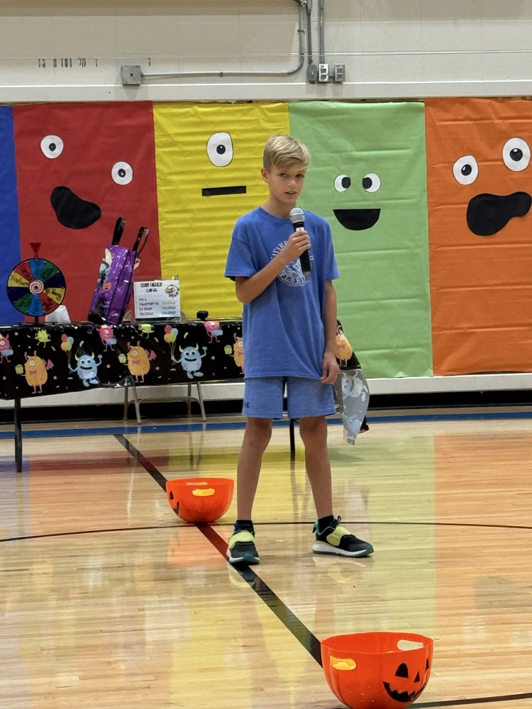 A young boy in a blue shirt sings into the microphone in an elementary school gym.