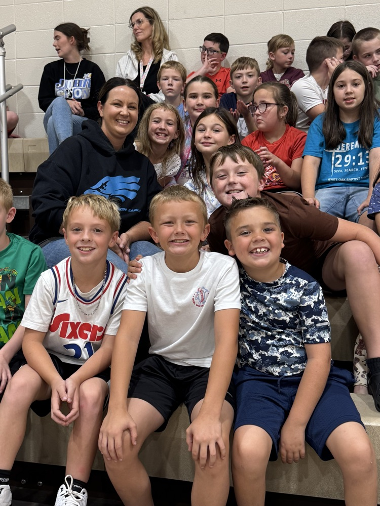 A large group of students surrounding their teacher smile while sitting on gym bleachers.