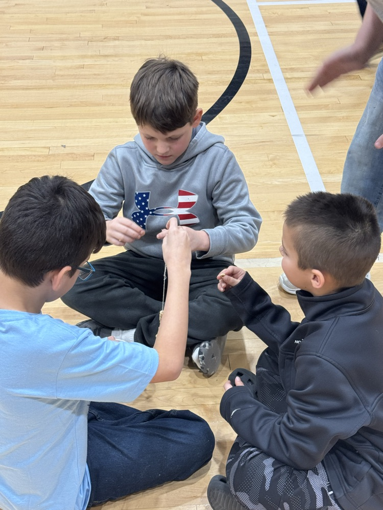 Three boys sit on the gym floor playing a game in which they have to put Cheerios on a piece of yarn in 100 seconds.