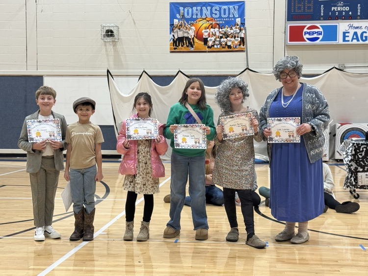 Five students in one adult stand on the gym floor, smiling, and holding their certificates for best dressed, senior citizen. They are all dressed in costume.
