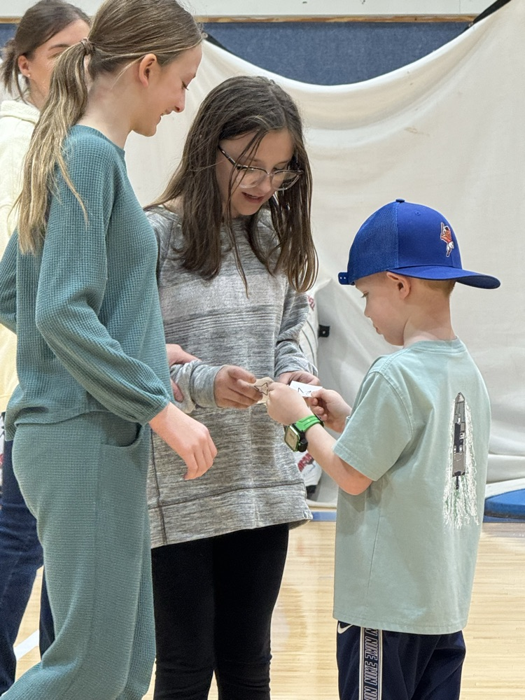 In the gym, two older girls help a younger boy in a backward blue baseball cap count paper stars that he collected as part of a game.
