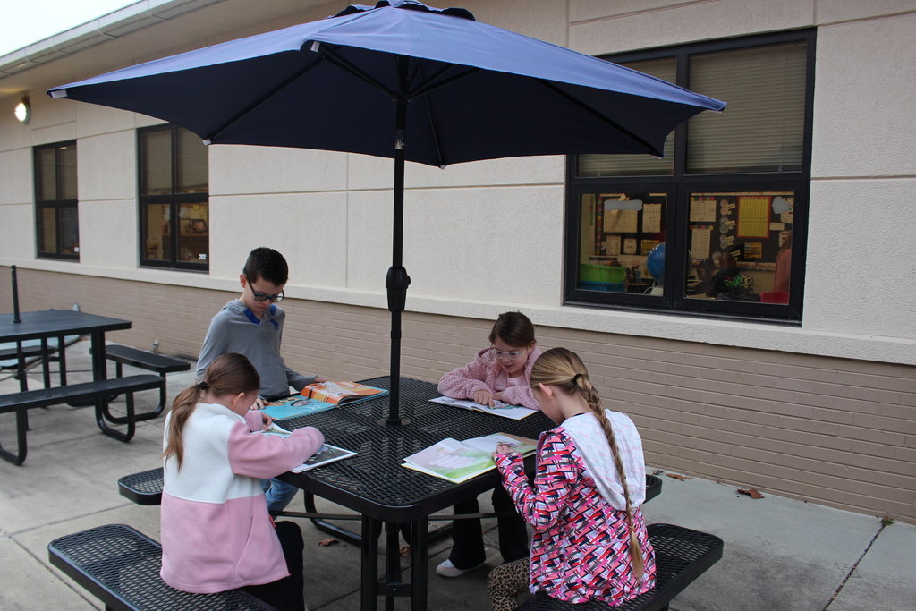 Elementary students sitting on their school patio at picnic table with umbrella reading