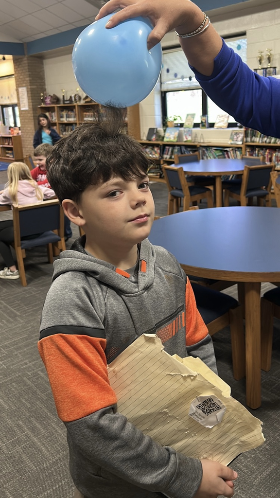 A boy in a gray and orange shirt looks toward the camera as someone standing outside the photo rubs his hair with a blue balloon