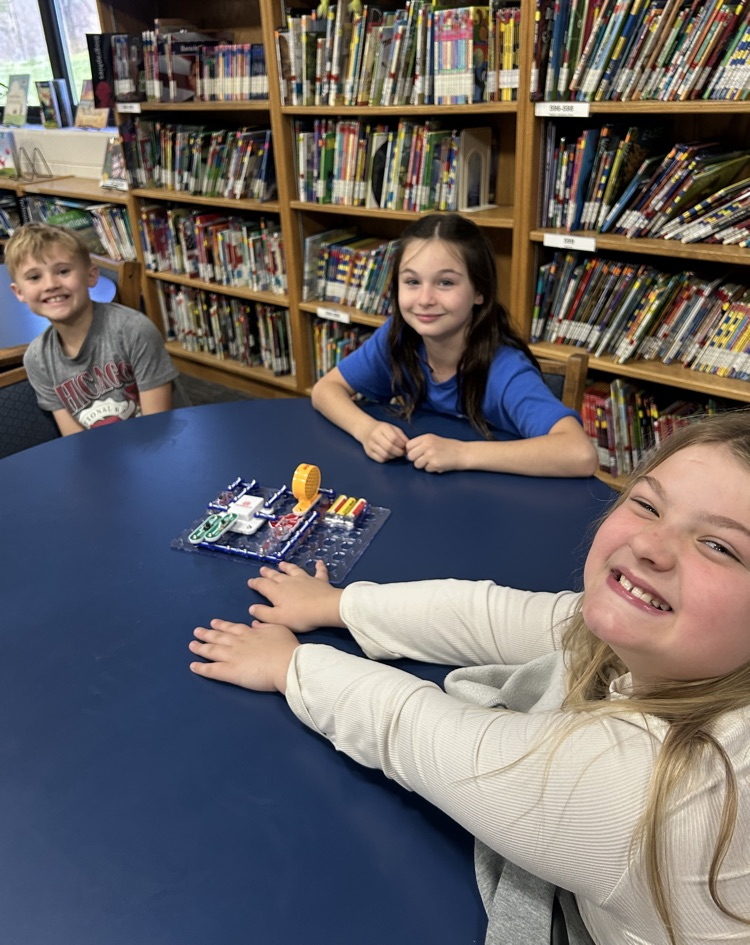 Three children smile for the camera as they sit around a round, blue table experimenting with a snap circuit kit