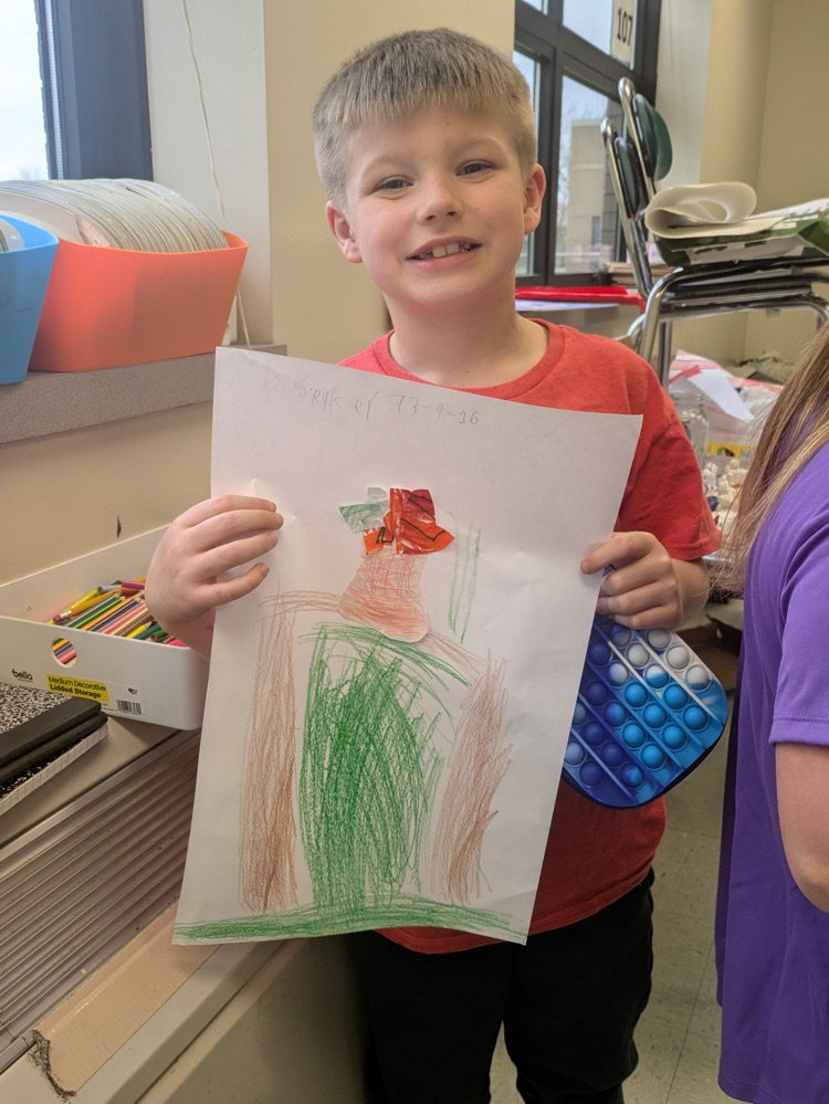 A boy smiles and holds up an art piece of a vase of flowers on a table  