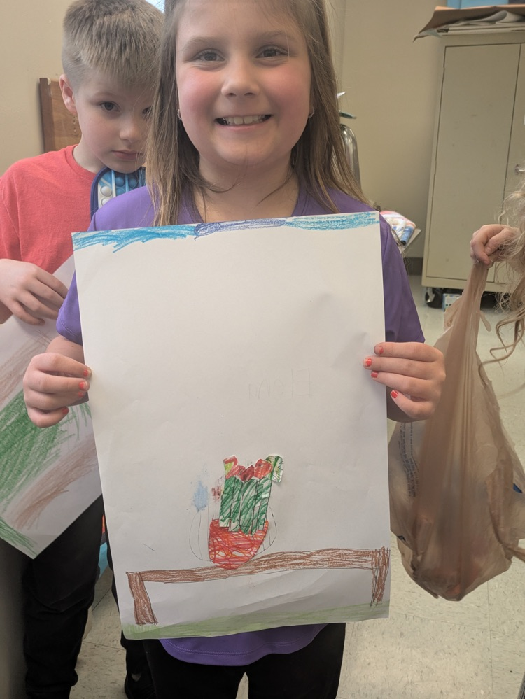 A girl smiles and holds up an art piece of a vase of flowers on a table. 