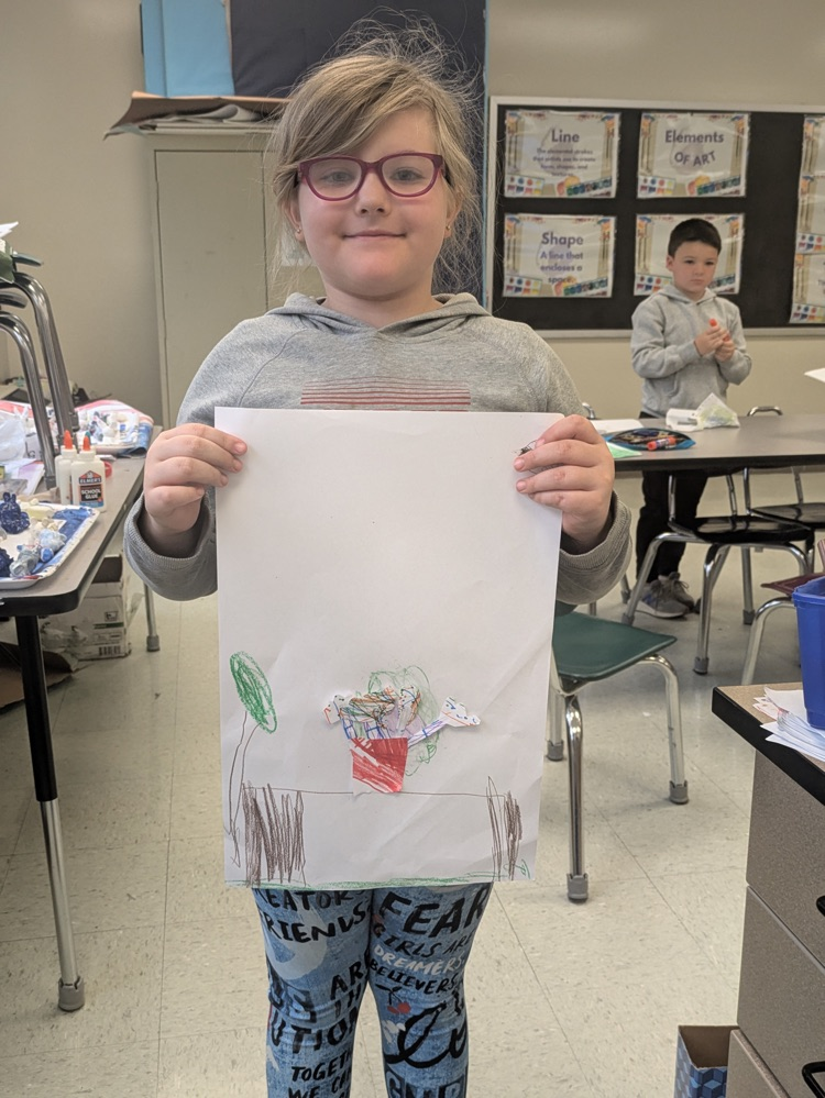 A girl smiles and holds up an art piece of a vase of flowers on a table.