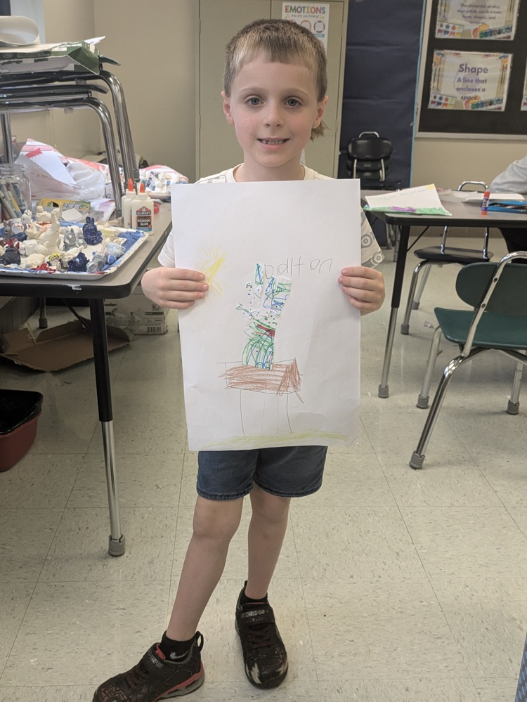 A boy smiles and holds up an art piece of a vase of flowers on a table