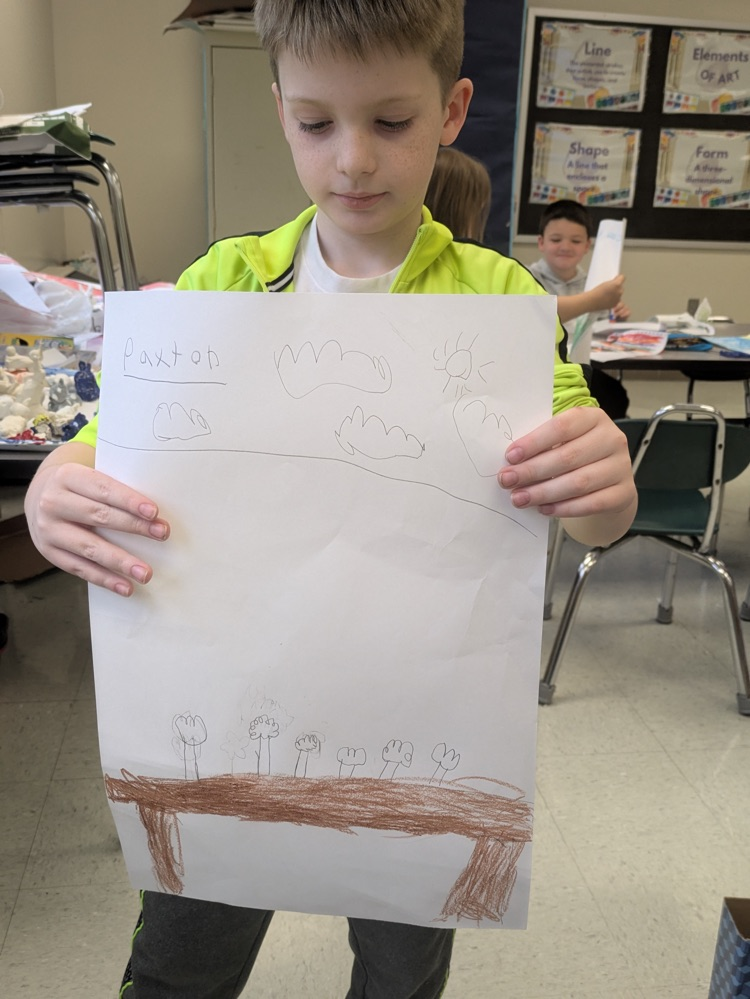 A boy smiles and holds up an art piece of a vase of flowers on a table