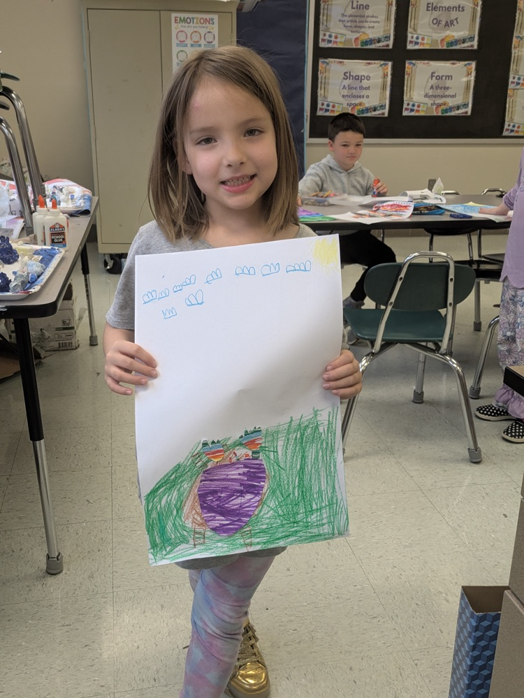 A girl smiles and holds up an art piece of a vase of flowers on a table