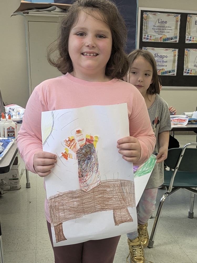 A girl smiles and holds up an art piece of a vase of flowers on a table