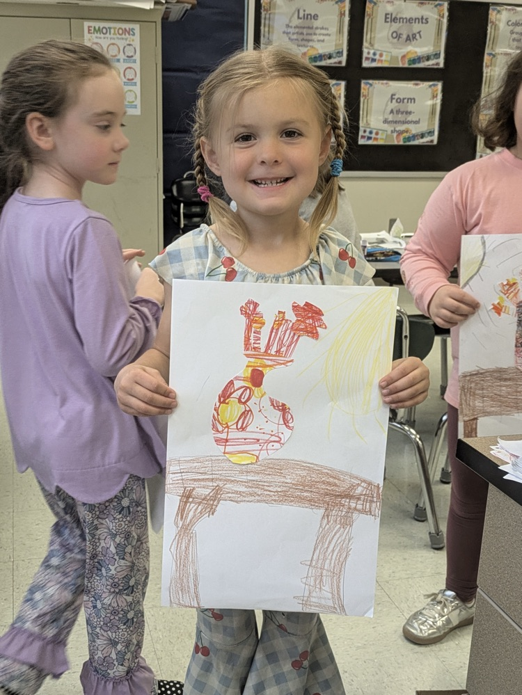 A girl smiles and holds up an art piece of a vase of flowers on a table