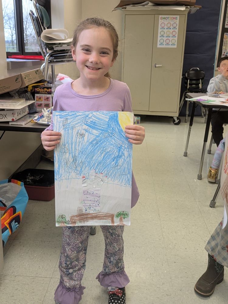A girl smiles and holds up an art piece of a vase of flowers on a table